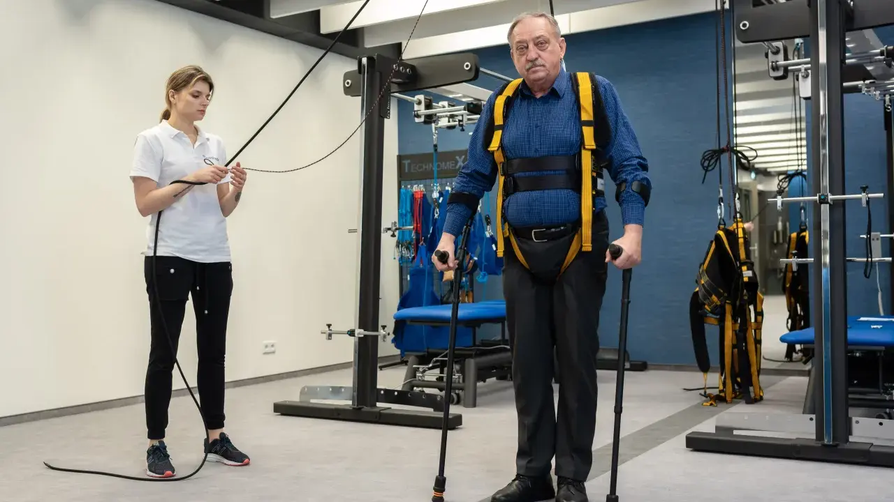 Starszy mężczyzna w specjalistycznej uprzęży wykonuje ćwiczenia rehabilitacyjne z pomocą fizjoterapeutki.