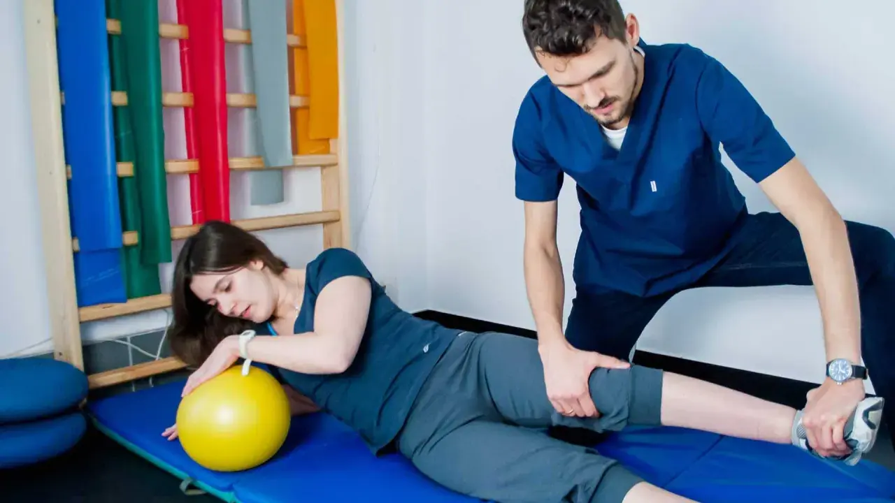 Fizjoterapeuta pomaga pacjentce w ćwiczeniach PNF. Kobieta leży na macie z żółtą piłką, a mężczyzna delikatnie naciska na jej nogę.