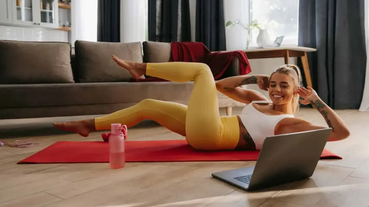 Uśmiechnięta kobieta wykonuje ćwiczenia w domu na macie, patrząc na laptopa.