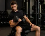 Mężczyzna w czarnym T-shircie wykonuje trening oporowy, podnosząc hantle. Skupiony na ćwiczeniu bicepsów.