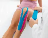 Fizjoterapeuta aplikuje plastry taping na kolano pacjentki, poprawiając stabilizację i wsparcie.