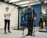 Starszy mężczyzna w specjalistycznej uprzęży wykonuje ćwiczenia rehabilitacyjne z pomocą fizjoterapeutki.