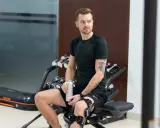 Mężczyzna w ortezie na kolanie, przygotowujący się do ćwiczeń rehabilitacyjnych łąkotki. Trzyma butelkę z wodą.
