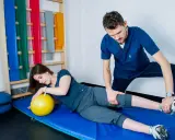 Fizjoterapeuta pomaga pacjentce w ćwiczeniach PNF. Kobieta leży na macie z żółtą piłką, a mężczyzna delikatnie naciska na jej nogę.