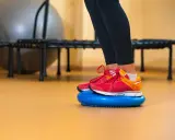 Nogi w butach sportowych stoją na niebieskiej poduszce sensorycznej, wykonując ćwiczenia równowagi.