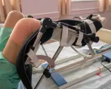 Pacjent w szpitalnym łóżku, z bandażem na kolanie, korzysta z urządzenia do rehabilitacji po endoprotezie kolana.