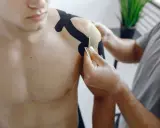 Fizjoterapeuta aplikuje taśmy kinezjotaping na bark mężczyzny. To część rehabilitacji barku po operacji, wspierająca ćwiczenia.