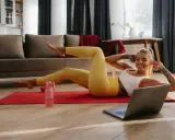 Uśmiechnięta kobieta wykonuje ćwiczenia w domu na macie, patrząc na laptopa.