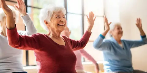 Uśmiechnięte seniorki wykonują ćwiczenia przeciwzakrzepowe, unosząc ręce w górę.