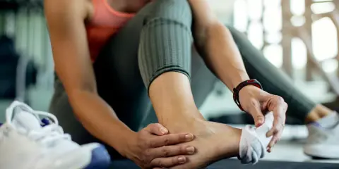 Kobieta z bólem po treningu, trzymająca się za stopę z powodu naderwanego więzadła.