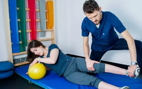 Fizjoterapeuta pomaga pacjentce w ćwiczeniach PNF. Kobieta leży na macie z żółtą piłką, a mężczyzna delikatnie naciska na jej nogę.
