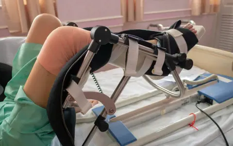 Pacjent w szpitalnym łóżku, z bandażem na kolanie, korzysta z urządzenia do rehabilitacji po endoprotezie kolana.