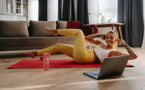 Uśmiechnięta kobieta wykonuje ćwiczenia w domu na macie, patrząc na laptopa.