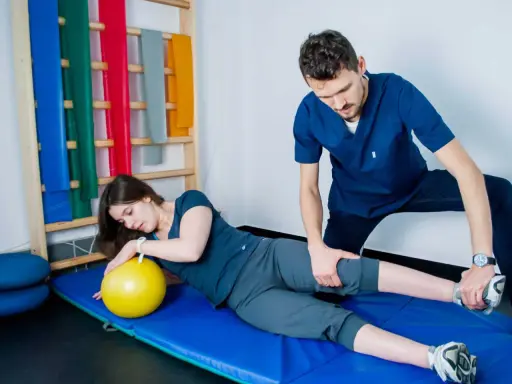 Fizjoterapeuta pomaga pacjentce w ćwiczeniach PNF. Kobieta leży na macie z żółtą piłką, a mężczyzna delikatnie naciska na jej nogę.