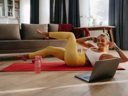 Uśmiechnięta kobieta wykonuje ćwiczenia w domu na macie, patrząc na laptopa.
