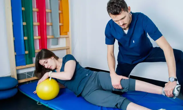 Fizjoterapeuta pomaga pacjentce w ćwiczeniach PNF. Kobieta leży na macie z żółtą piłką, a mężczyzna delikatnie naciska na jej nogę.