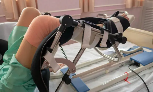 Pacjent w szpitalnym łóżku, z bandażem na kolanie, korzysta z urządzenia do rehabilitacji po endoprotezie kolana.