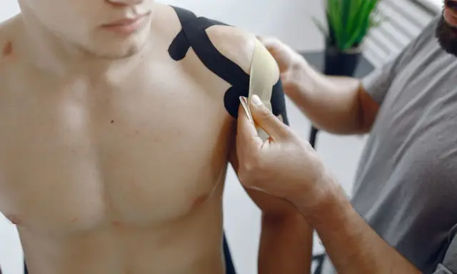 Fizjoterapeuta aplikuje taśmy kinezjotaping na bark mężczyzny. To część rehabilitacji barku po operacji, wspierająca ćwiczenia.
