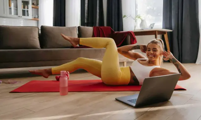 Uśmiechnięta kobieta wykonuje ćwiczenia w domu na macie, patrząc na laptopa.