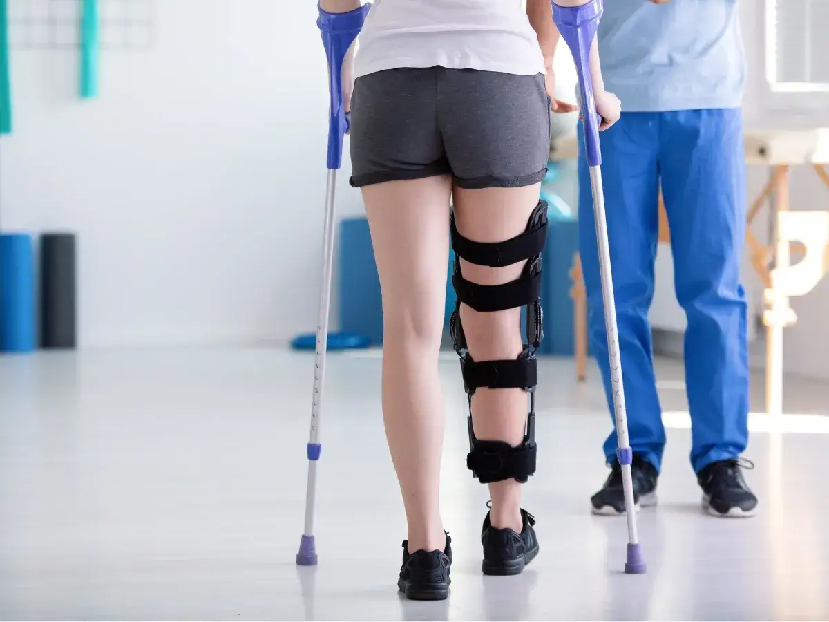 Rehabilitacja po zdjęciu gipsu: pacjentka z ortezą na nodze i kulami, wspierana przez fizjoterapeutę.