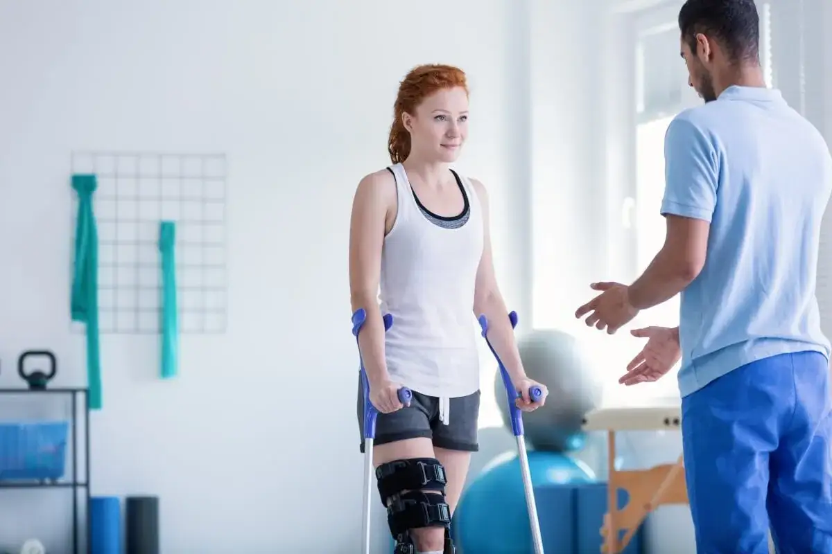 Fizjoterapeuta pomaga kobiecie z kulami i ortezą na nodze. Długość rehabilitacji zależy od postępów.