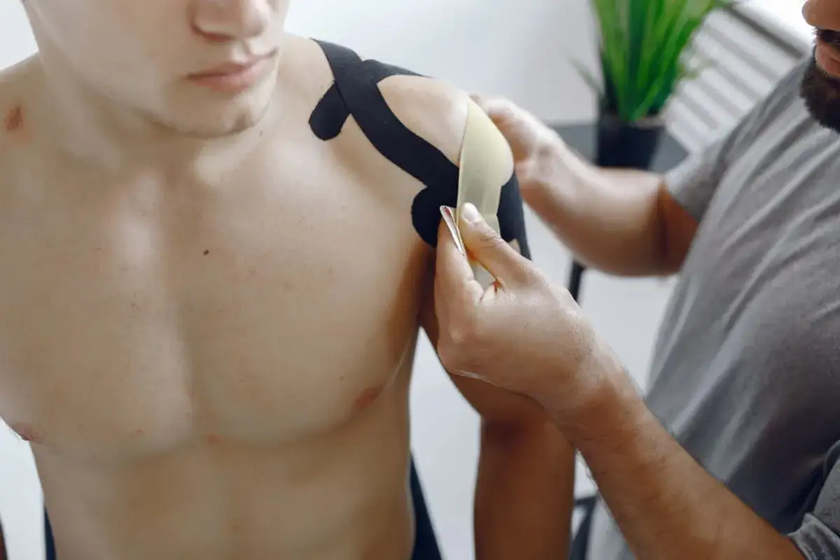Fizjoterapeuta zakłada taśmy kinezjotaping na bark mężczyzny. To część rehabilitacji barku po operacji, wspierająca ćwiczenia.